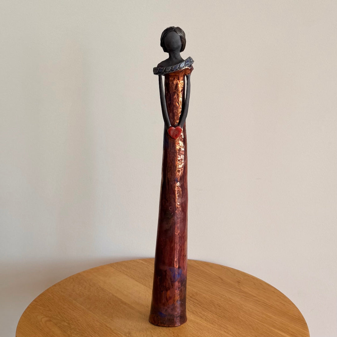 Raku fired ceramic figurine holding a red heart on top of a wooden stand against a plain background
