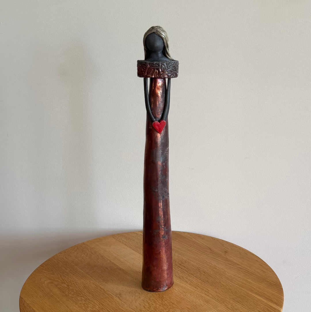 Raku fired ceramic figurine holding a red heart on top of a wooden stand against a plain background