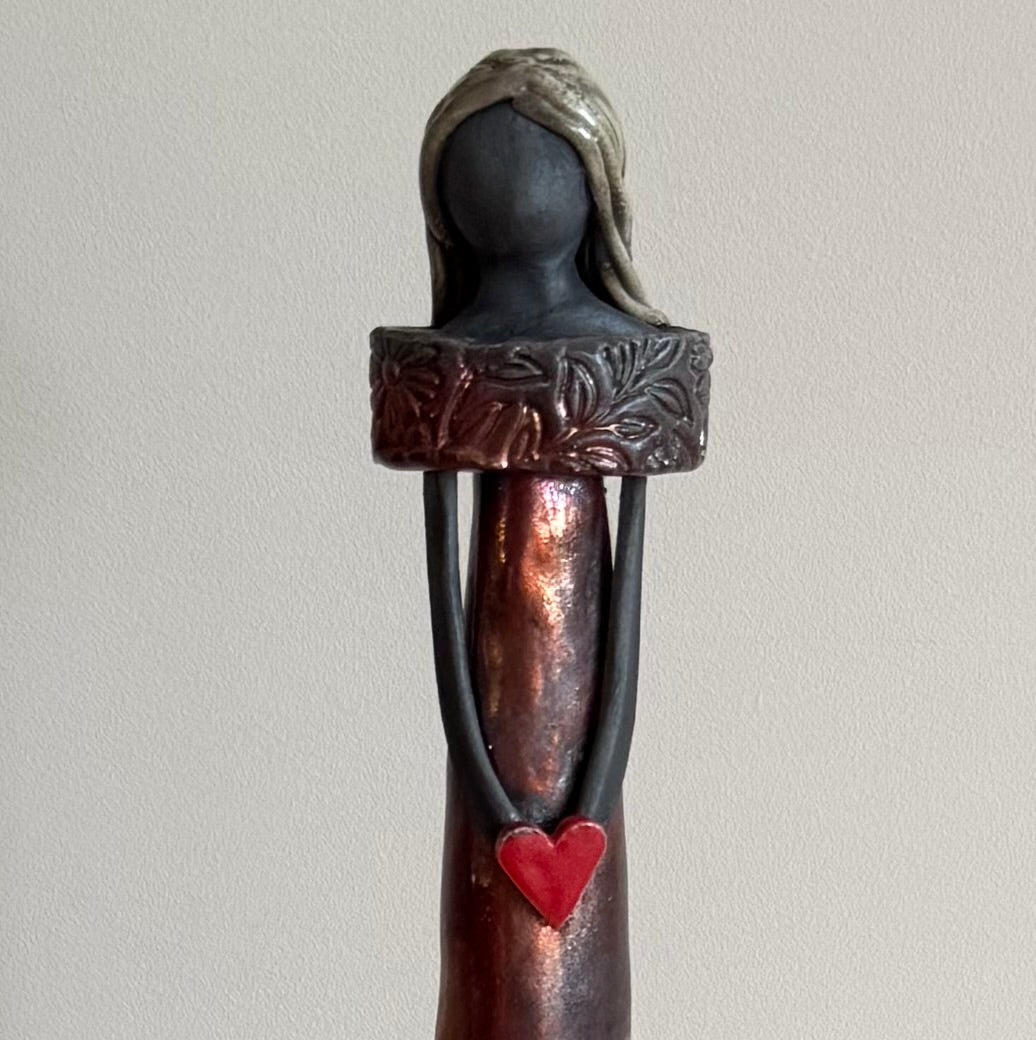 Close up half view of a raku fired ceramic figurine holding a red heart on top of a wooden stand against a plain background