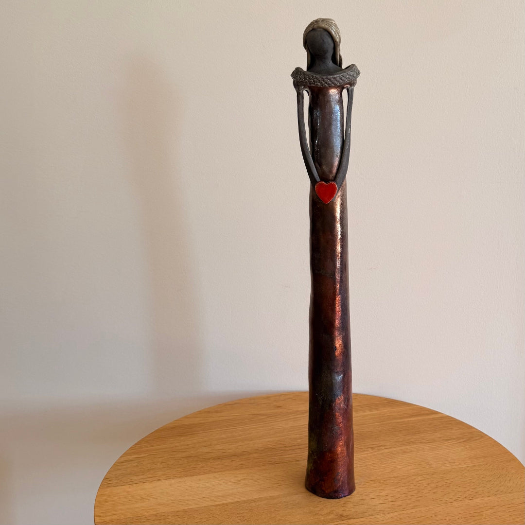 Ceramic sculpture of a figure with a heart on a wooden stand against a plain background