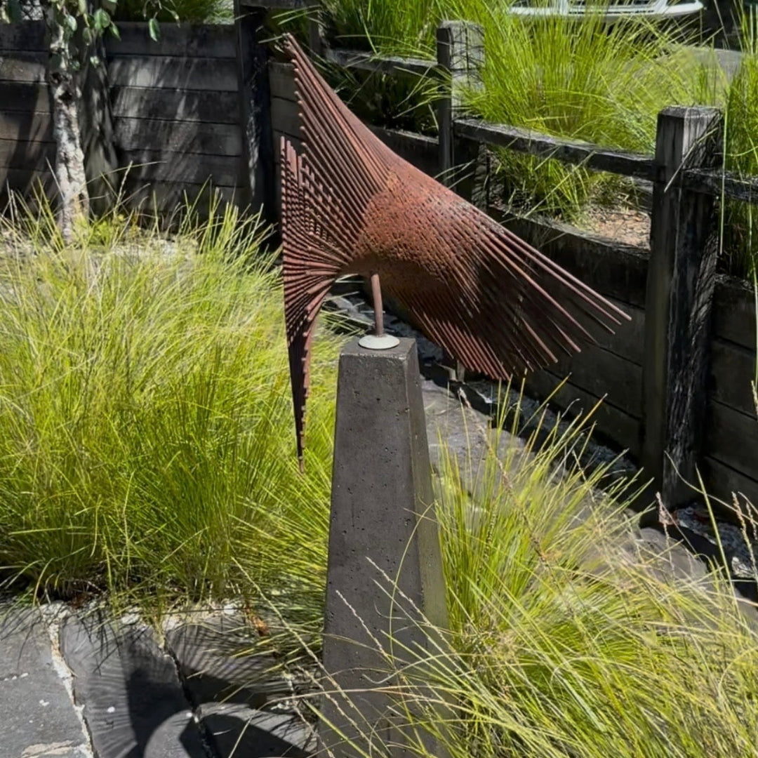 Decorative metal sculpture on a pedestal in a garden setting with grass and wooden fence.