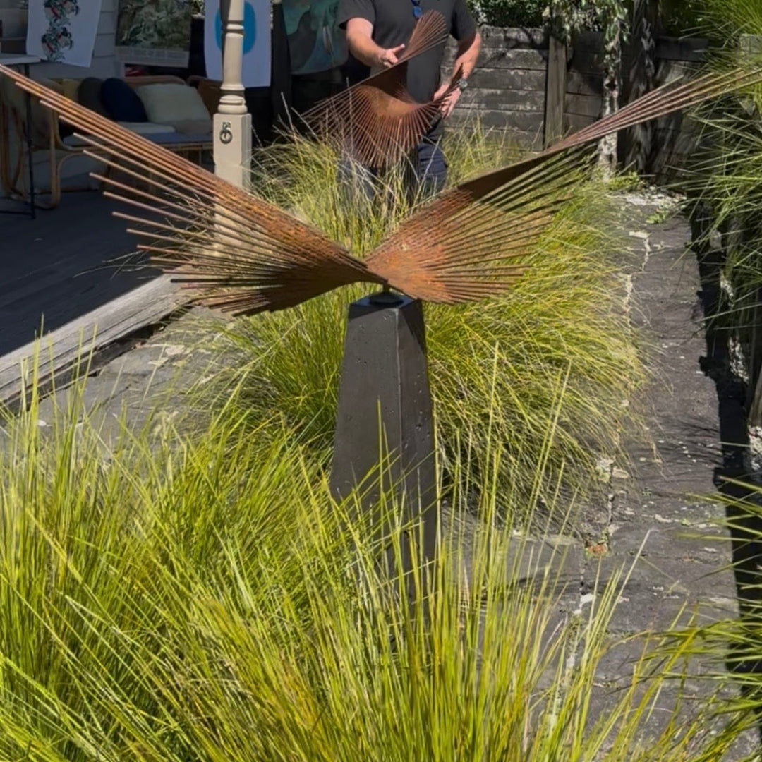 Metal sculpture resembling a bird in flight surrounded by tall grasses.