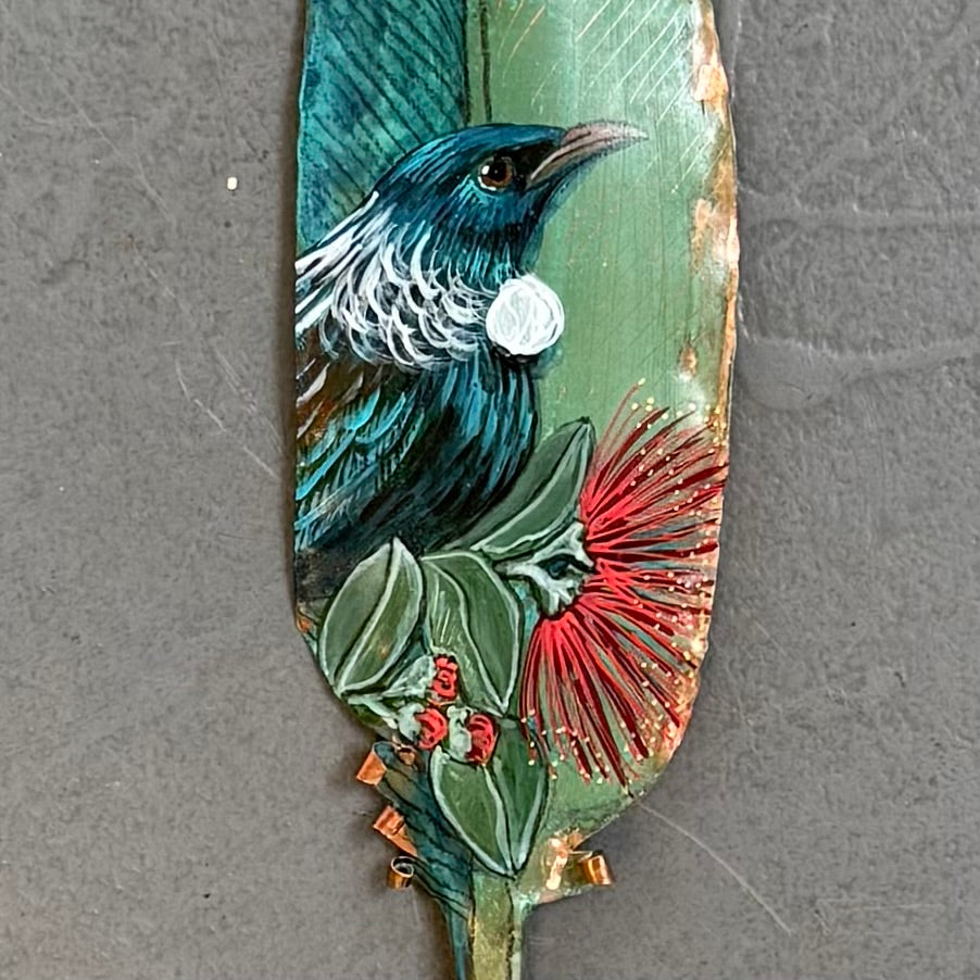 Close up of a decorative copper feather with a painted Tui bird & flowers on a gray background