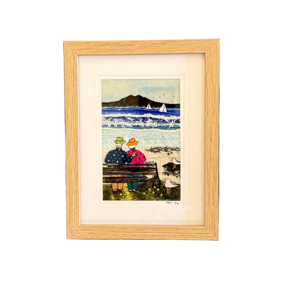 Framed textile artwork of two people on a beach with sailboats and Rangitoto in the background.
