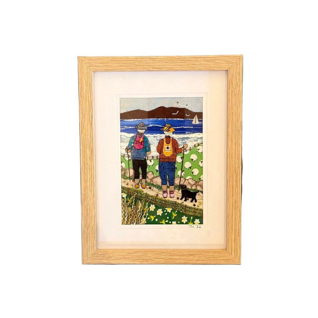 Framed textile artwork of two people with a dog on a grassy field, mounted on a white wall.