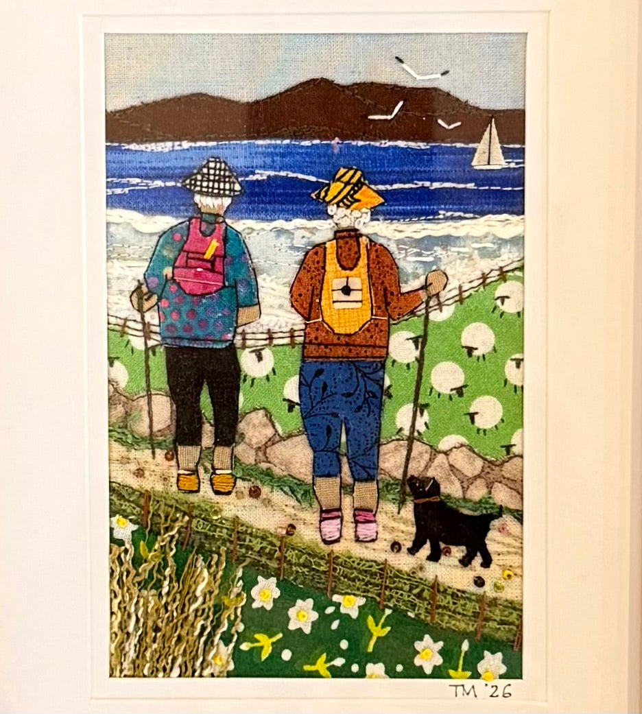 Textile artwork of two people walking with a dog by a beachside with mountains and sheep in the background.