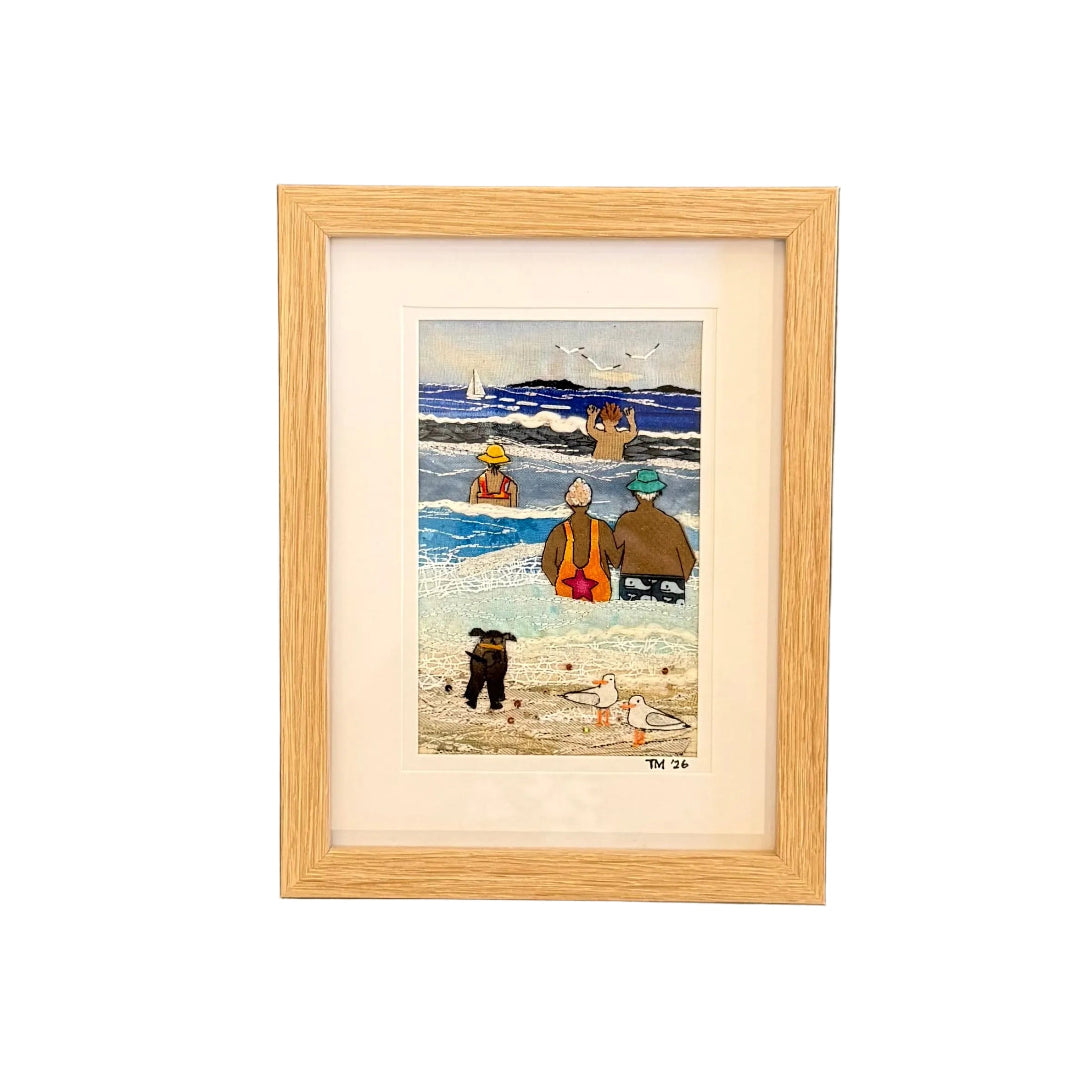 Framed textile artwork of two people swimming at the beach with dog.