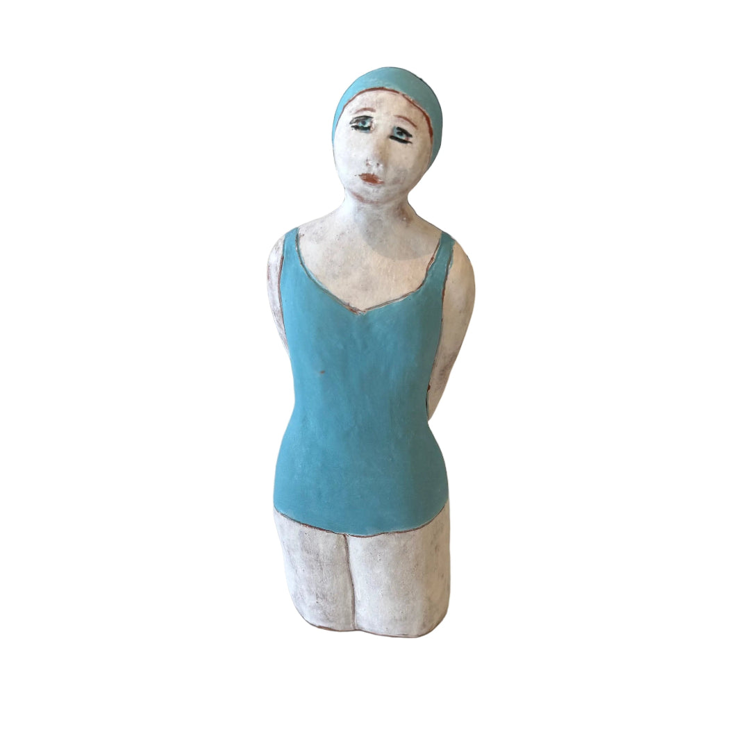 Ceramic sculpture of a Swimmer in blue swimsuit & cap with hands behind her back on a white background