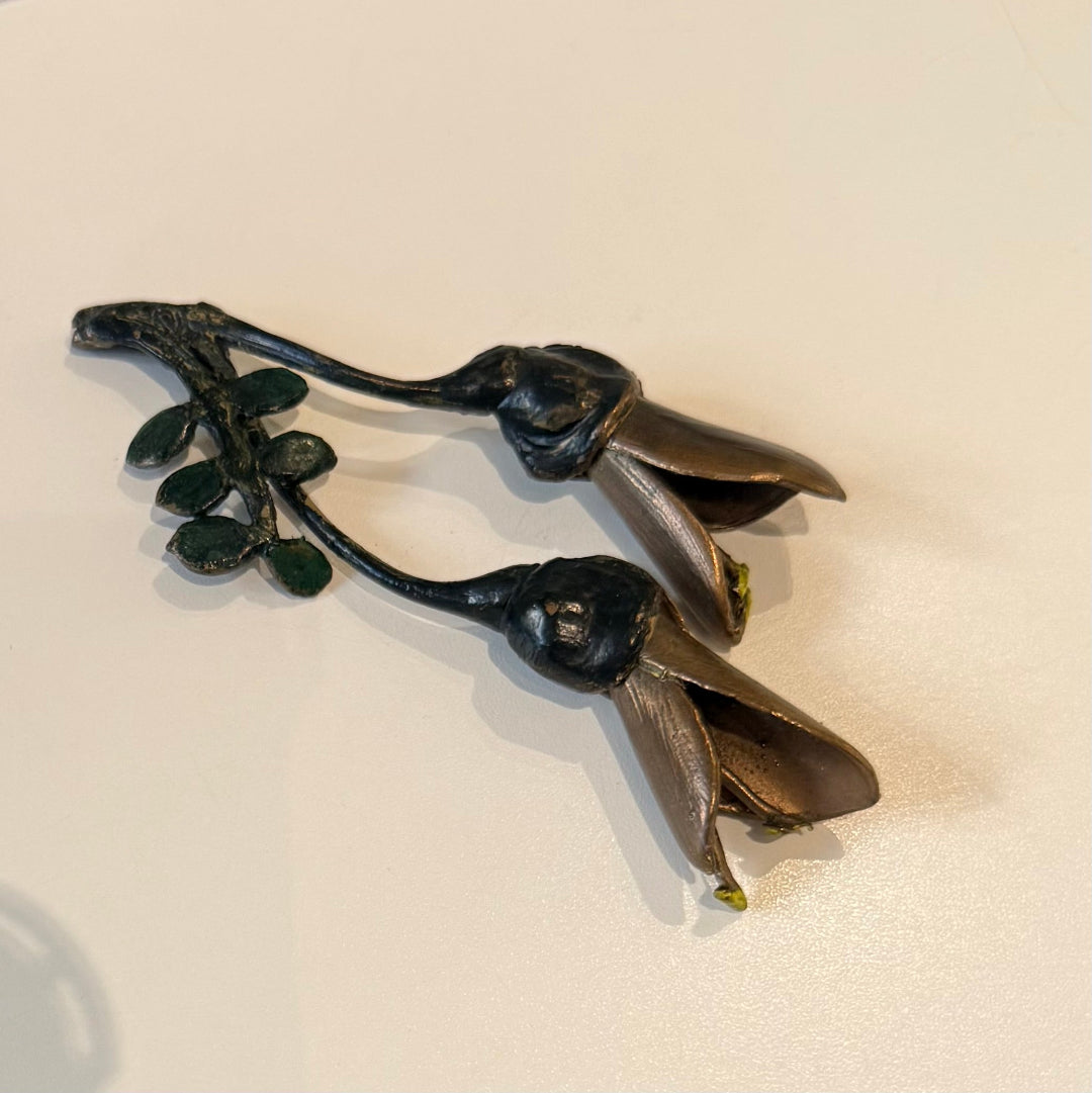 A pair of miniature painted bronze Kowhai flowers with visible seed pods, placed on a plain background.