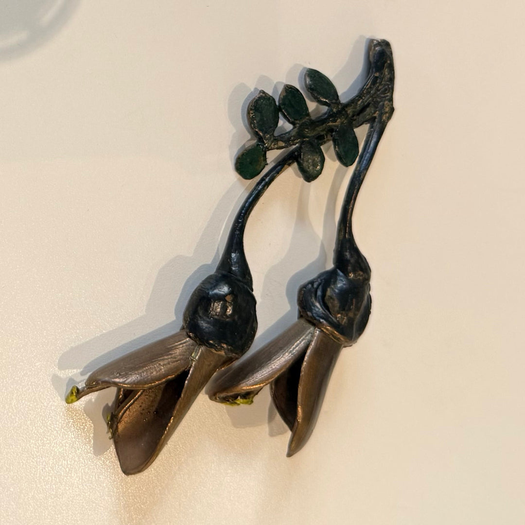 A pair of miniature painted bronze Kowhai flowers with visible seed pods, placed on a plain background.