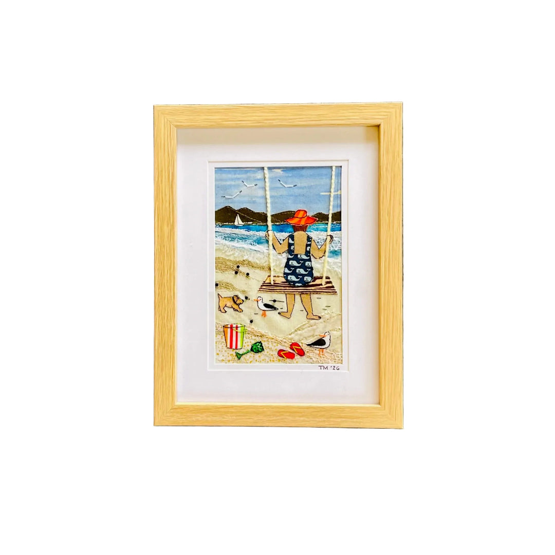 Framed textile art depicting a woman sitting on a beach swing, viewed from behind, with dogs, seagulls, and the sea in the background. The print is matted and housed in a light natural wood frame, mounted on a white wall.