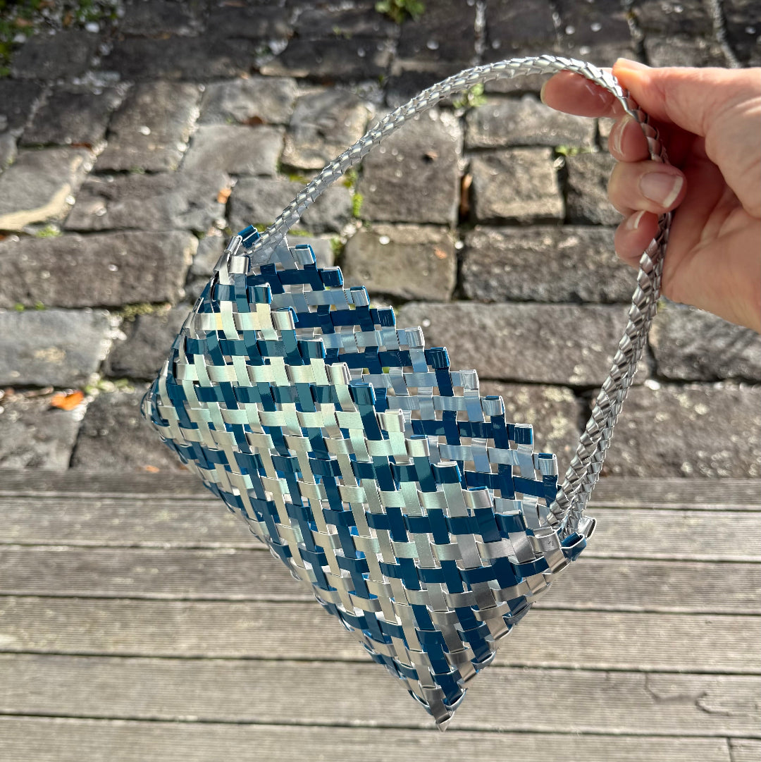Hand holding a woven blue and silver bag or Kete on a wooden surface with a brick background