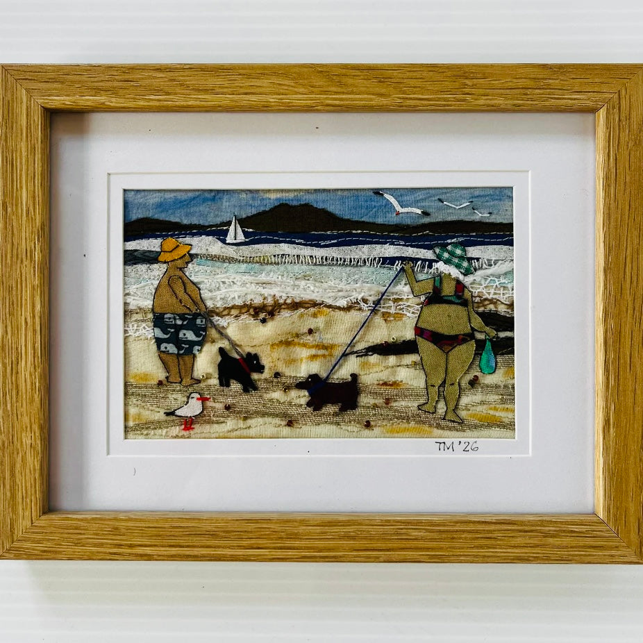 Framed artwork of a beach scene with people and dogs, featuring a sailboat and mountains in the background.