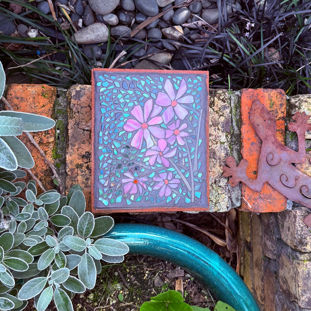Decorative paver with floral mosaic on a stone wall with plants and a garden hose.