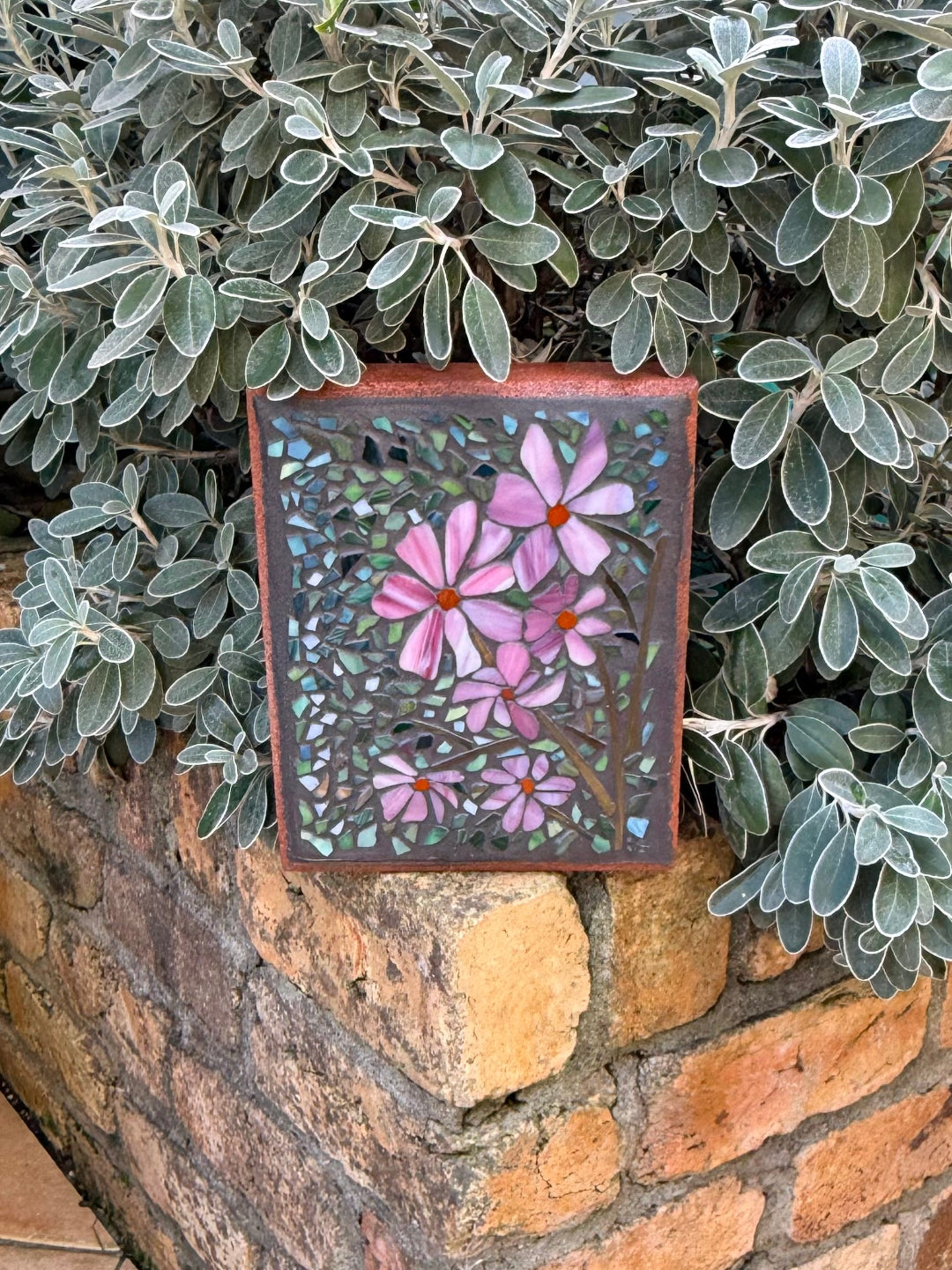 Decorative mosaic paver with pink flowers on a stone wall and green foliage background