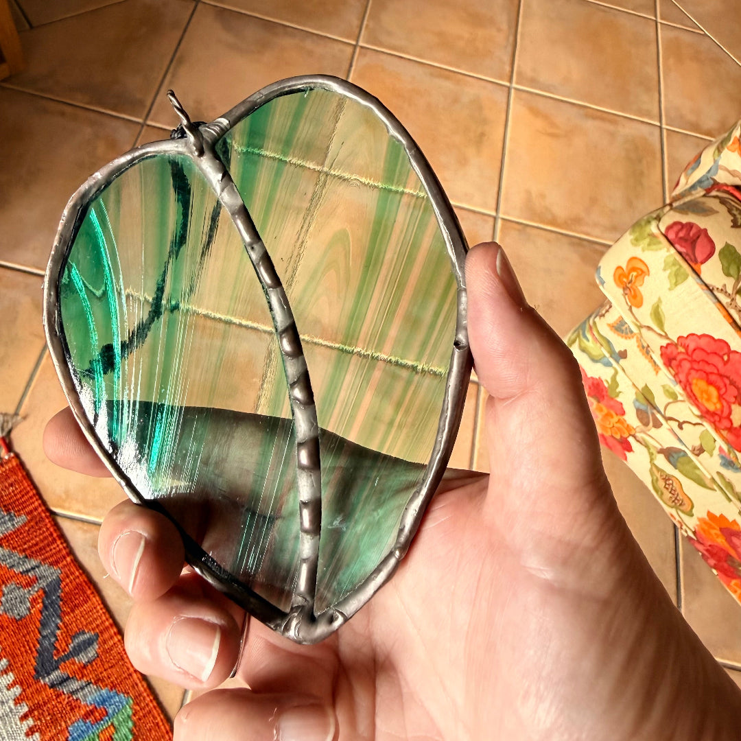 Hand holding a leaf-shaped stained glass wall hanging above a tiled floor.