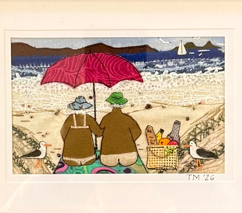 Textile artwork of two people under a red umbrella on a beach with a basket of food and seagulls.