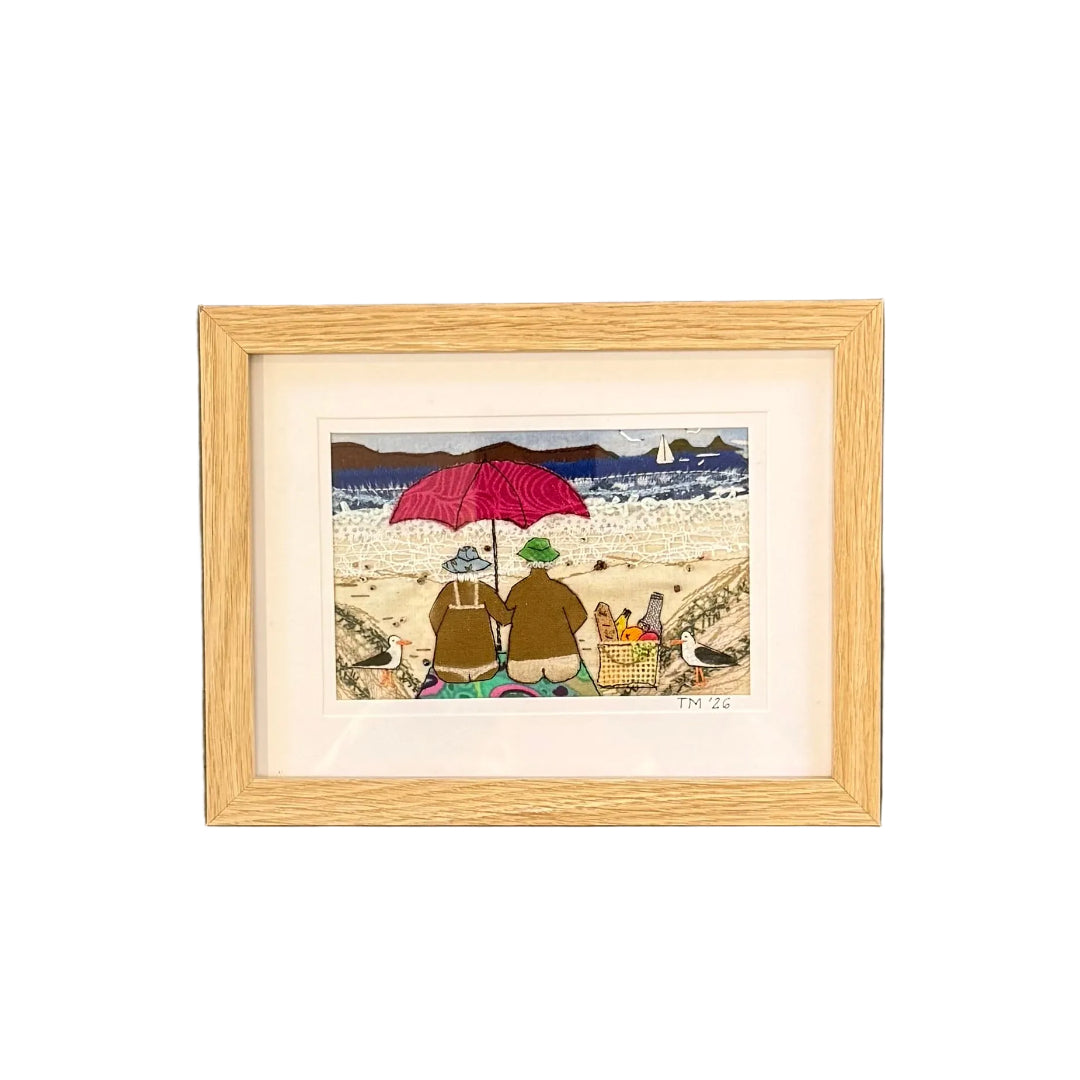 Framed textile artwork of two figures under a red umbrella on a beach, mounted on a white wall.