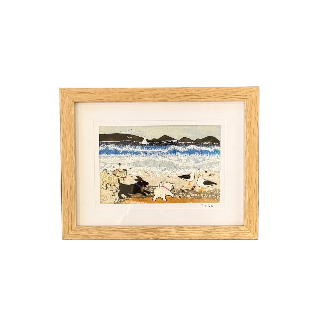 Framed textile artwork of 3 dogs at the beach on a white wall.