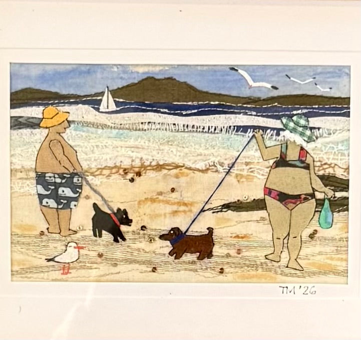 Textile artwork of a beach scene with two people and two dogs.