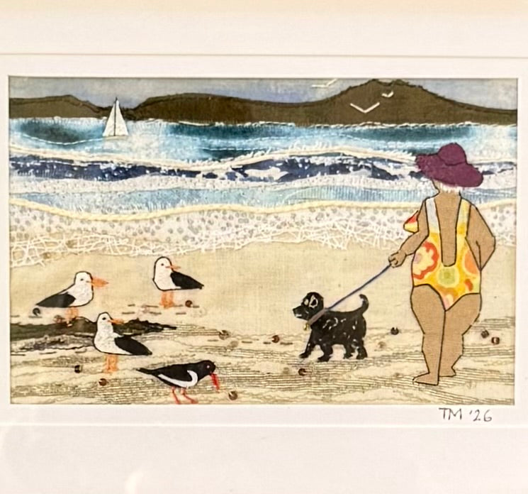Textile artwork of a person walking a dog on a beach with birds and sailboats in the background.