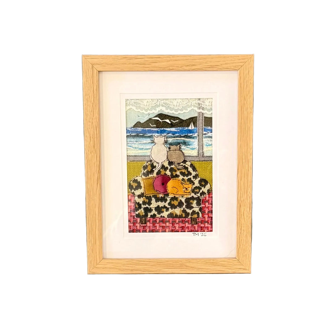 Textile artwork of 3 cats in a room looking at the ocean with a headland with a white wall background