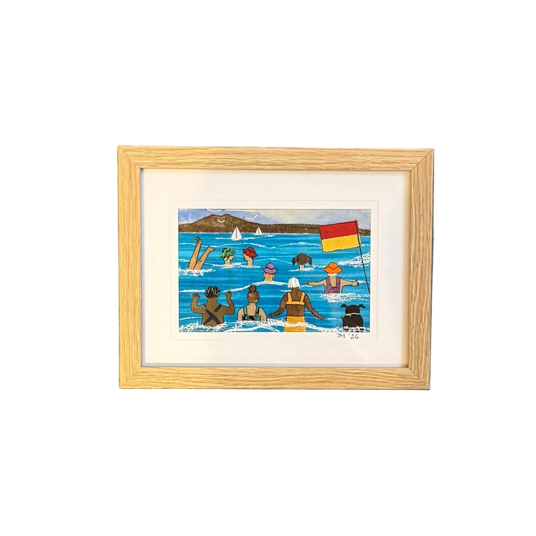 Framed textile artwork of a beach scene with people and flags on a white wall.