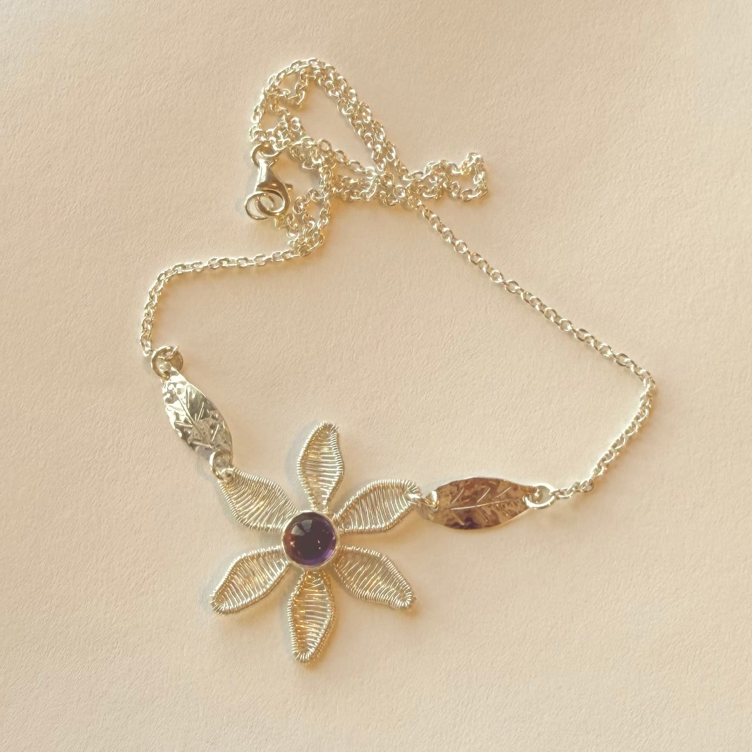Silver necklace with floral design and purple gemstone on a beige background