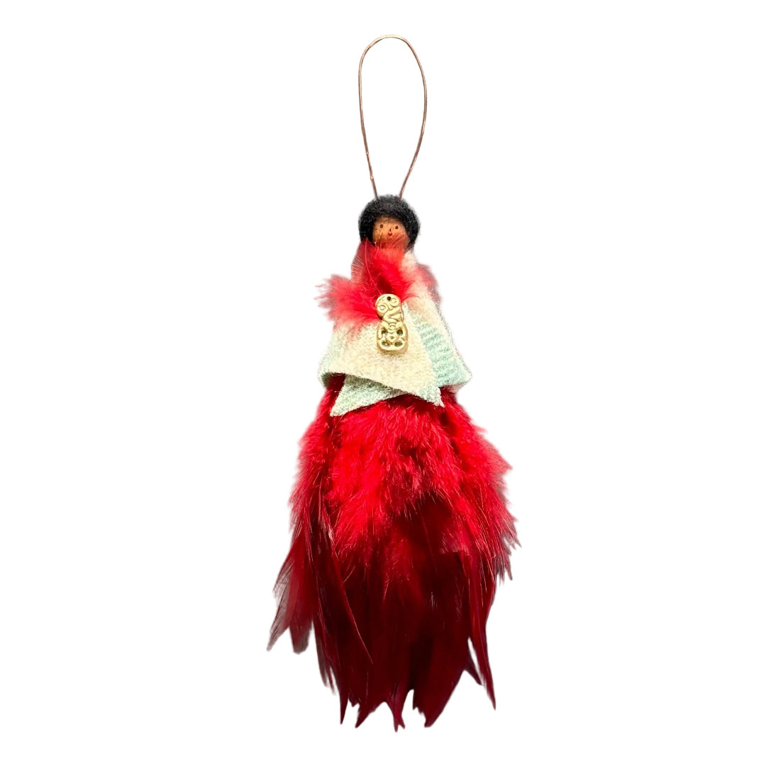 A decorative hanging ornament shaped like a small doll with a black hat, red feathered dress, pale green & cream fabric shawl, and a gold tiki ornament on the chest, suspended by a thin copper wire loop against a plain white background.