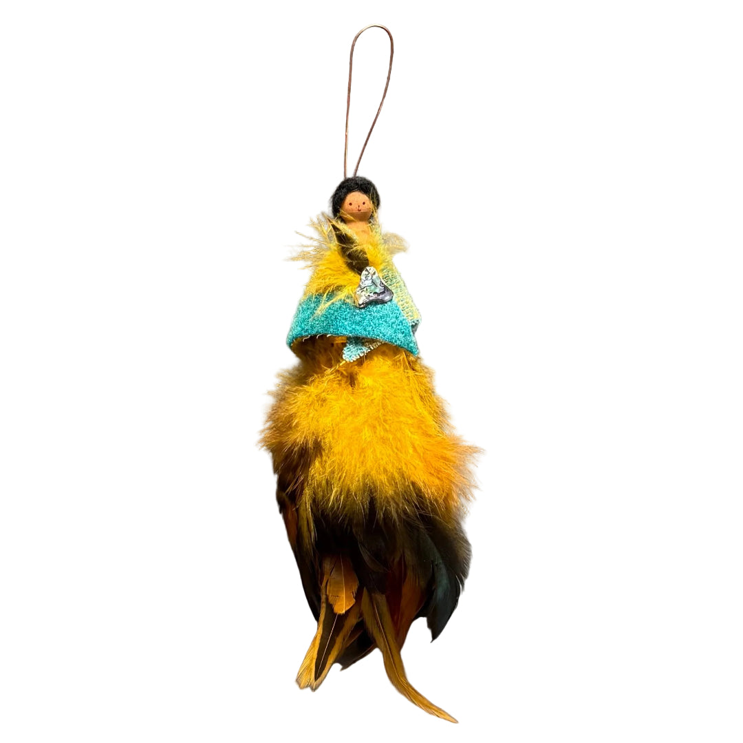 A handmade decorative hanging ornament featuring a small peg doll figure with black hair and a face, dressed in a turquoise felt shawl with a paua shell & feather brooch , and a fluffy yellow-and-brown feather skirt, suspended against a white background.