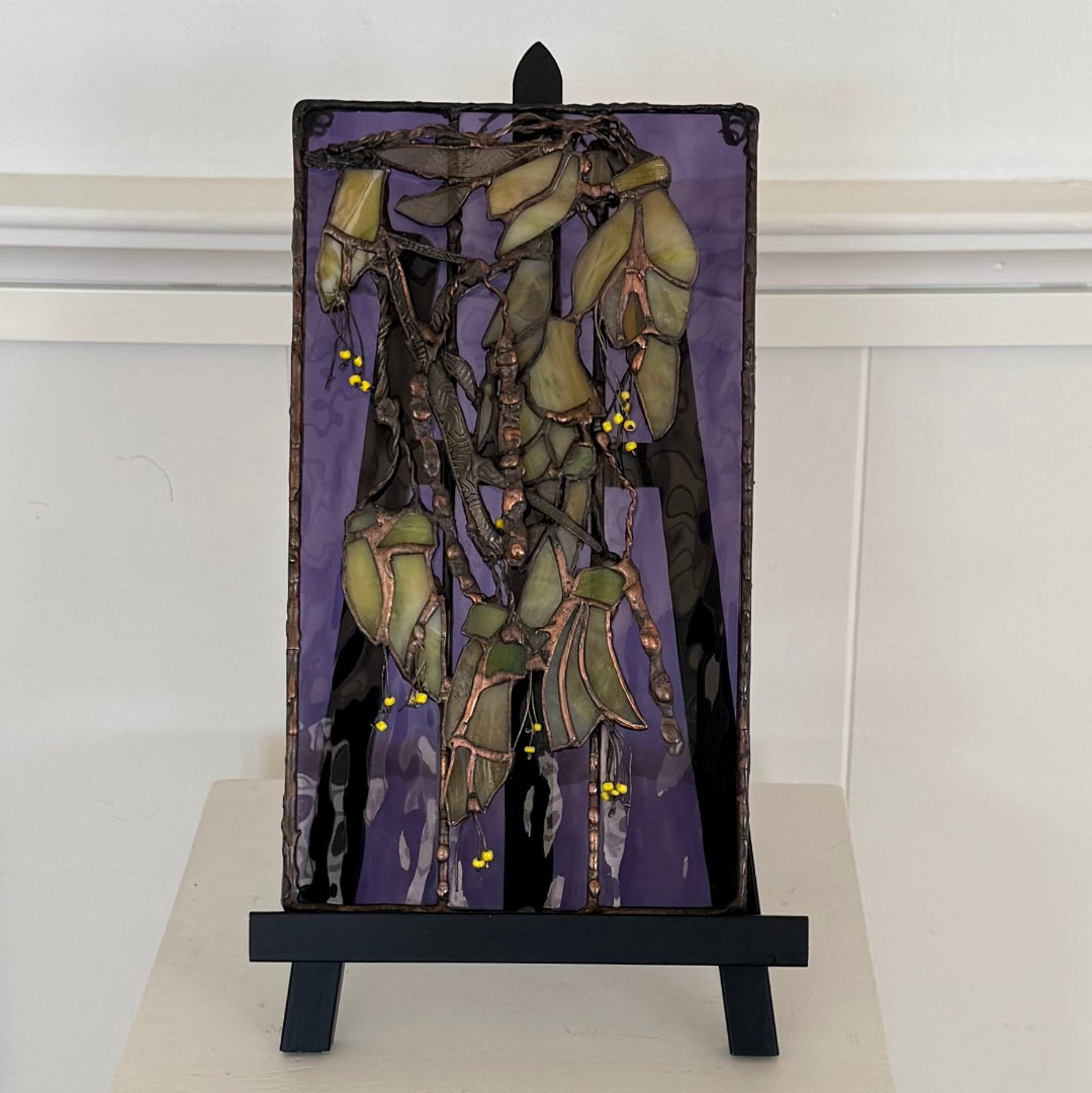 A rectangular stained glass artwork depicting stylized kowhai flowers, displayed on a black easel stand against a white paneled wall.