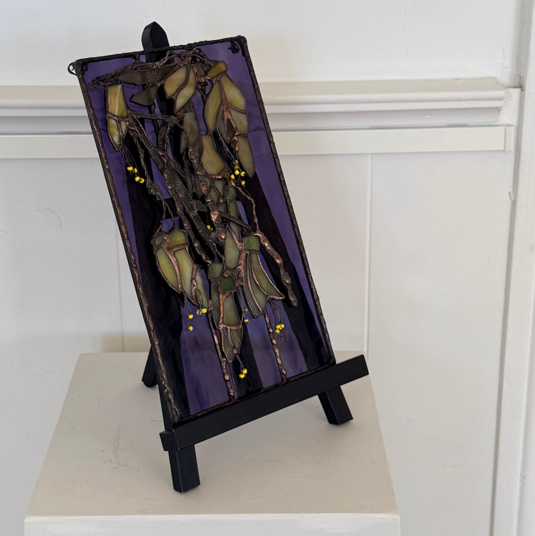 A stained glass art piece depicting hanging leaves and kowhai flowers in olive green, gold, and brown tones over a purple background, displayed on a black easel stand against a white paneled wall.