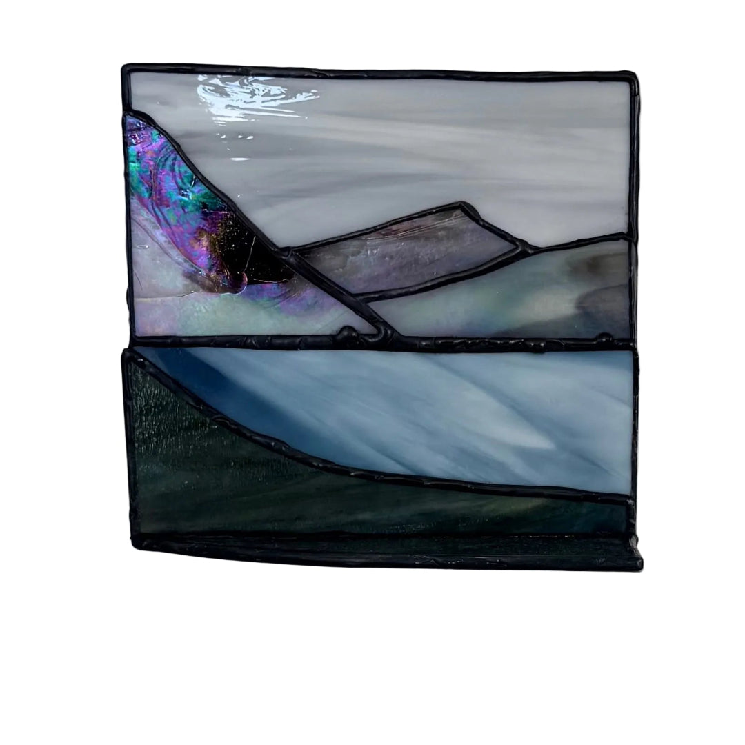 A rectangular stained glass panel depicting a landscape with mountains and flowing blue water, featuring a purple and iridescent section on the left, set against a plain light background.