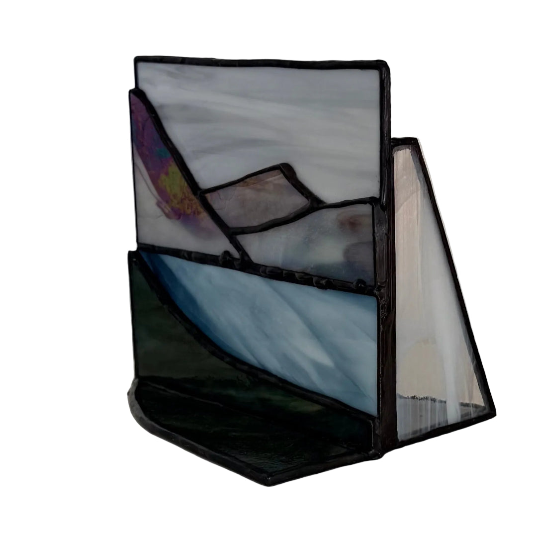 A handmade stained glass art piece depicting abstract mountain and landscape scenes, featuring a gray and white sky, blue water, and dark green foreground, held together by black solder.