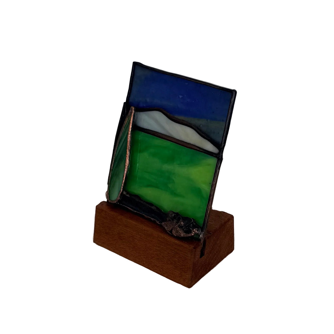 A small stained glass art panel with a geometric landscape design featuring blue, green, and white panels, mounted on a brown wooden base.