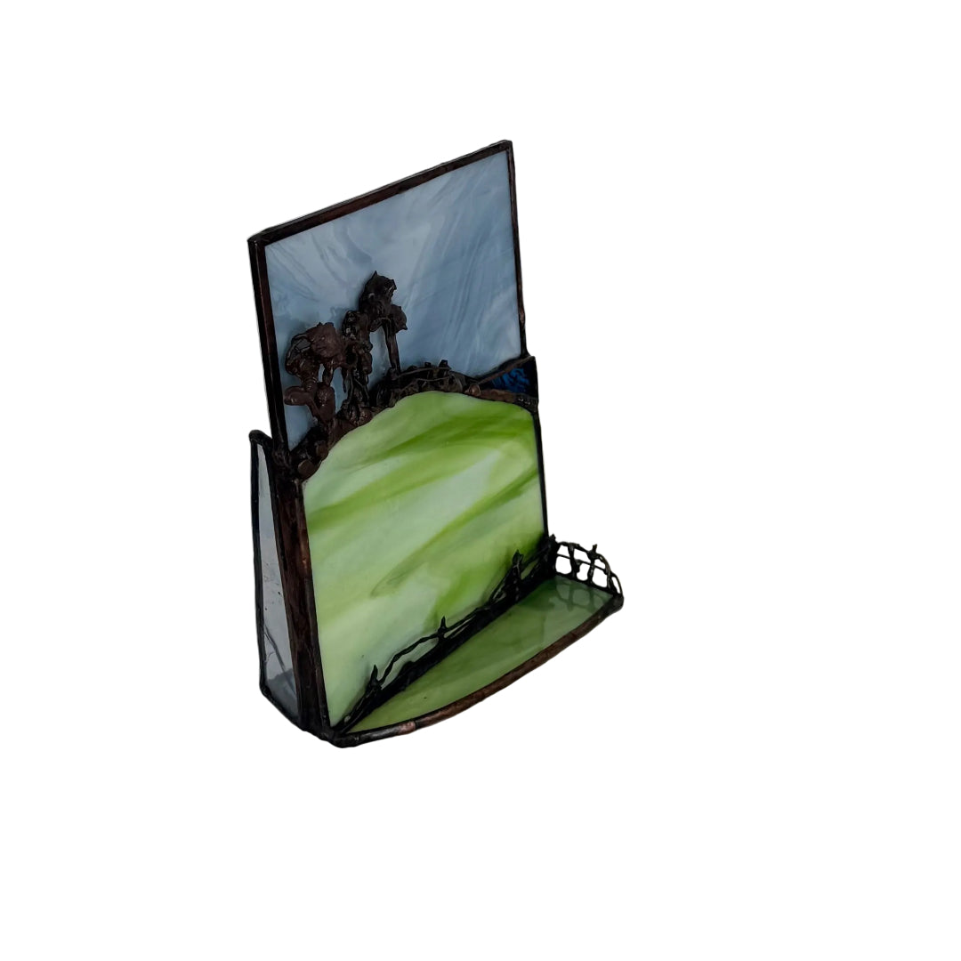 A small decorative frame with a landscape design, featuring green hills, dark brown trees, and a blue sky, displayed on a light-colored surface against a white paneled wall.