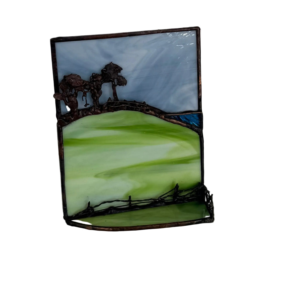 A stained glass landscape frame with green fields, dark metal tree silhouettes, and a light blue sky, displayed on a light-colored surface against a white wall.