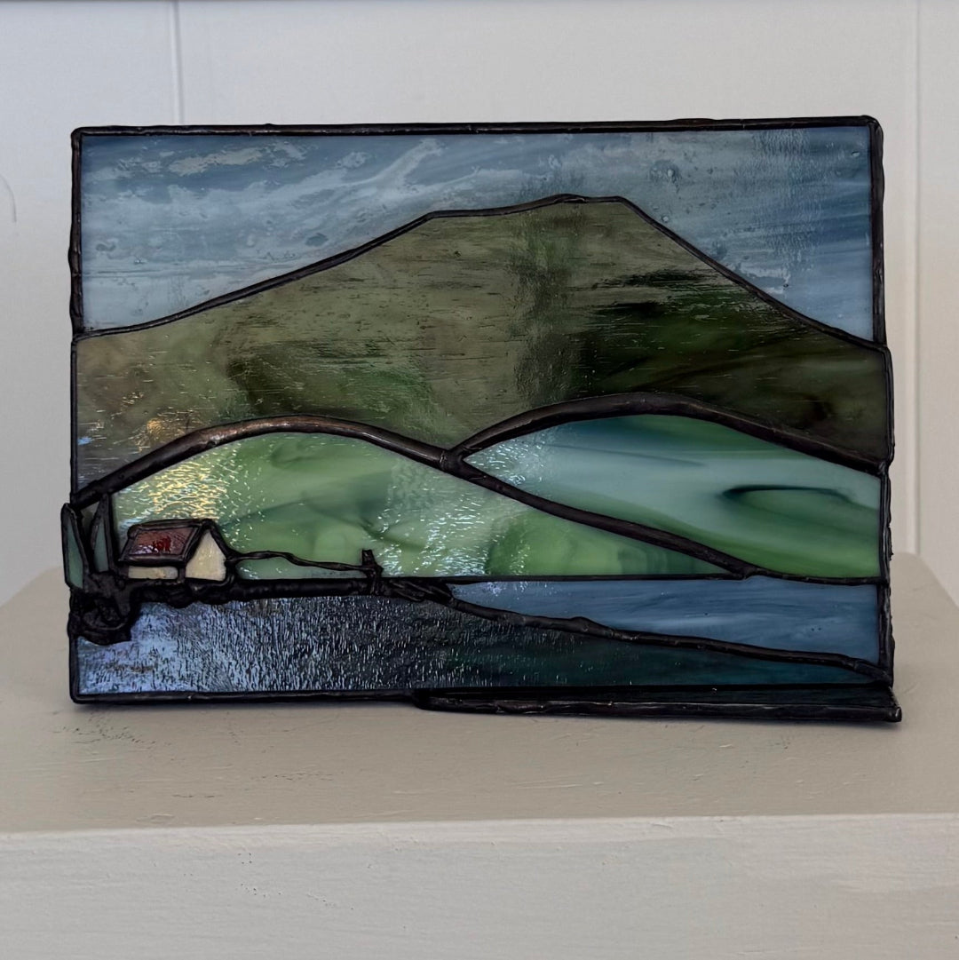 A rectangular stained glass panel depicting a mountain landscape with green hills, blue-gray sky and water, a small house, and dark lead outlines, displayed against a white wall and shelf.