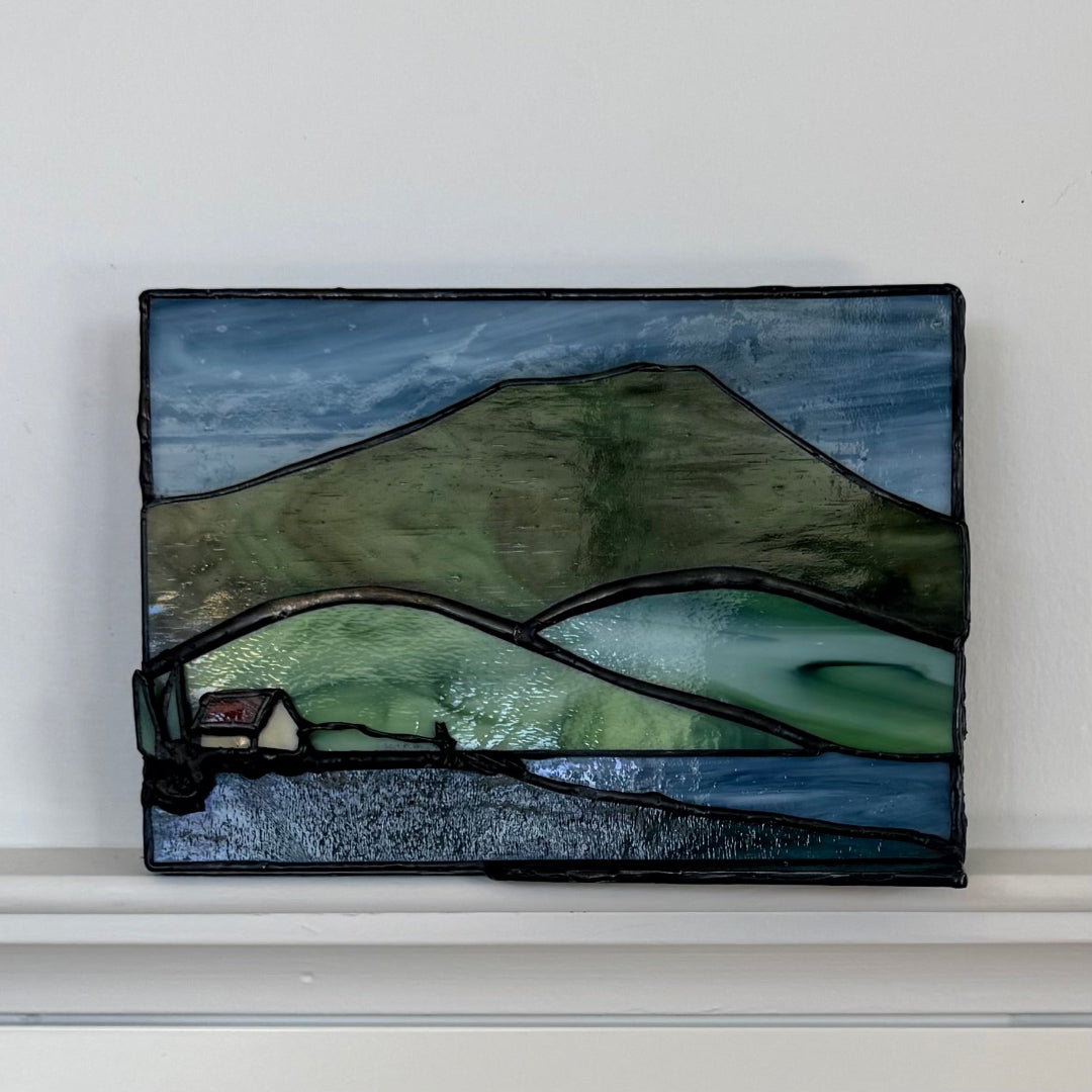 A rectangular stained glass art piece depicting a mountain landscape with green hills, blue sky, and water, mounted on a white wall above a white shelf.