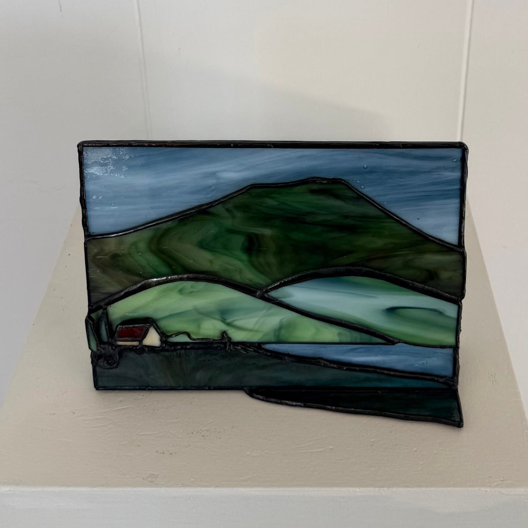 A rectangular stained glass panel depicting a mountain landscape with green and blue tones, resting on a light beige surface against a white wall.