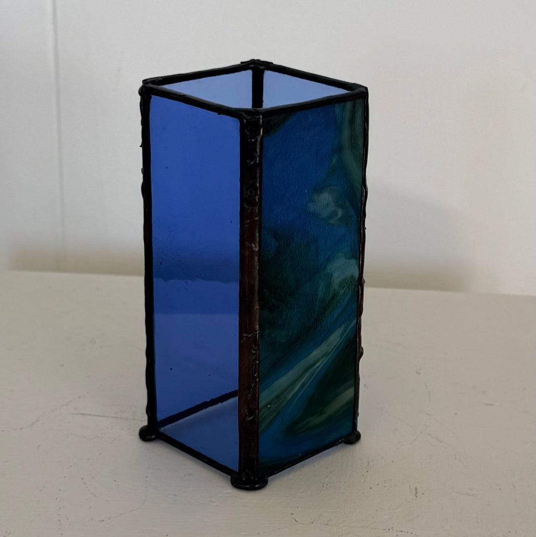 A small rectangular glass lantern or box with a black metal frame, featuring cobalt blue glass panels and one darker marbled blue-green panel, standing on a white surface against a white wall.