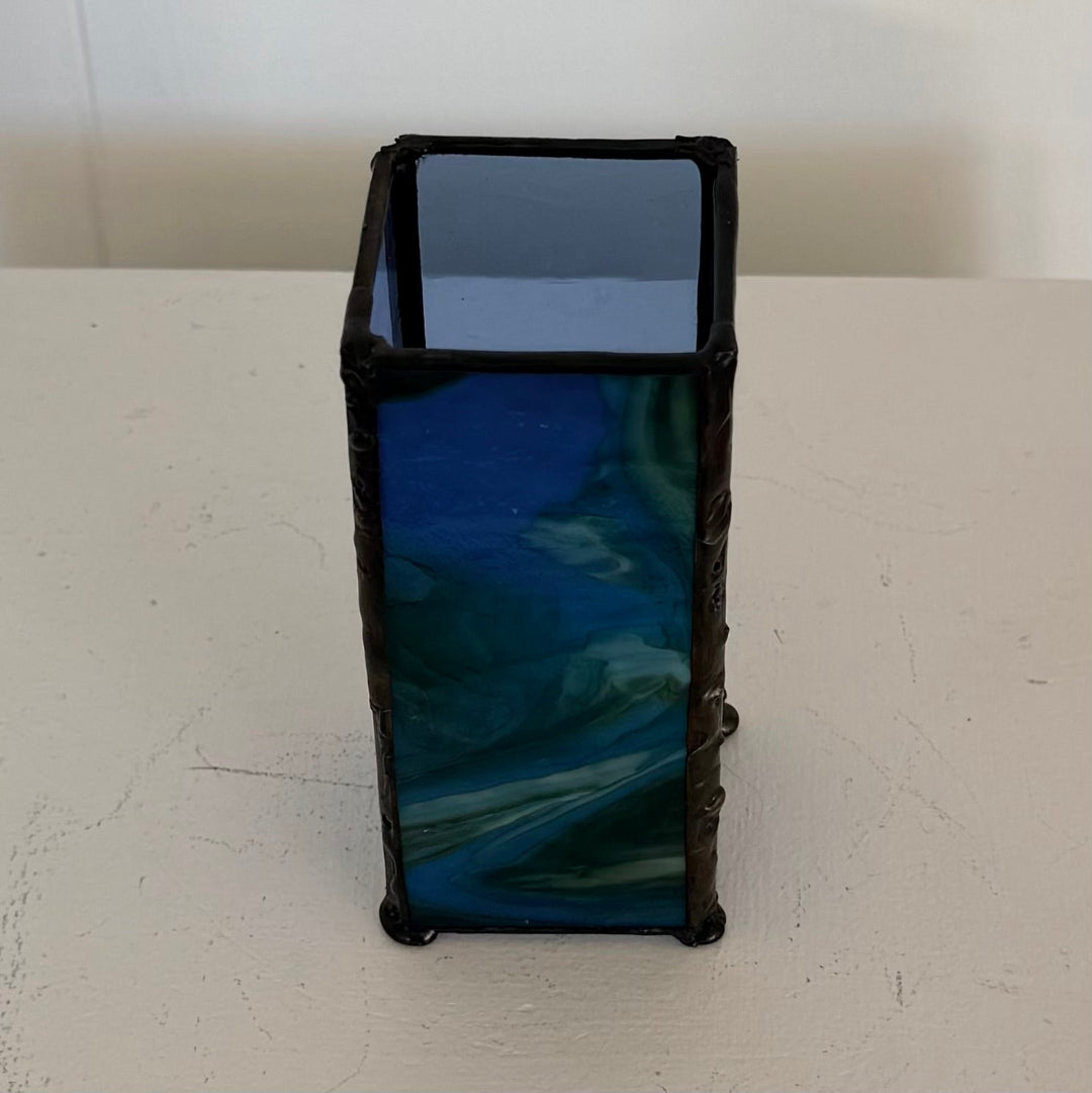 A rectangular glass vase or candle holder with a dark blue marbled pattern and a dark brown or black textured rim, standing on a white surface against a white wall.