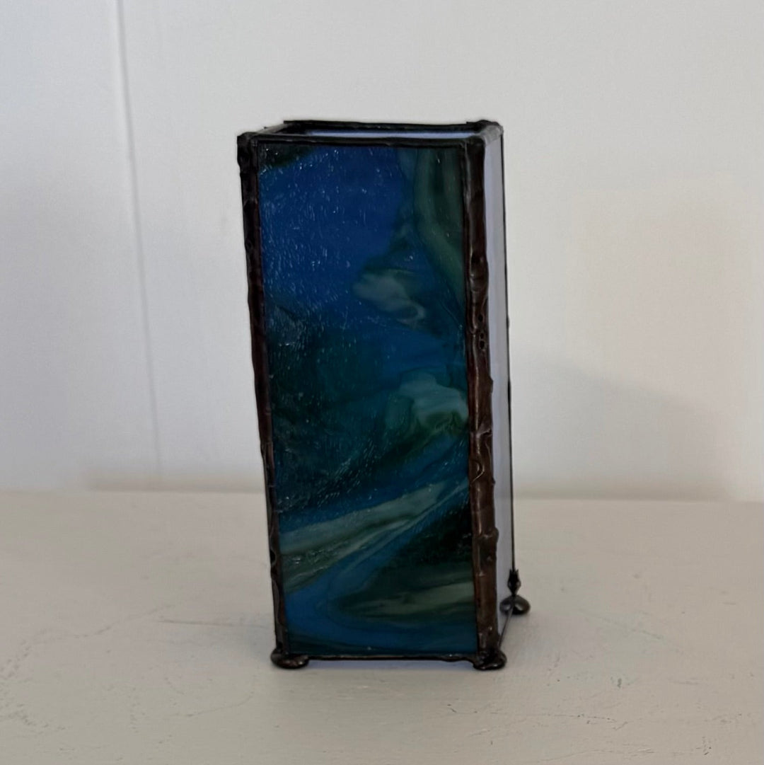 A rectangular glass vase with a dark blue and teal marbled pattern, framed by dark brown or black metallic edging, standing on a light-colored surface against a white wall.