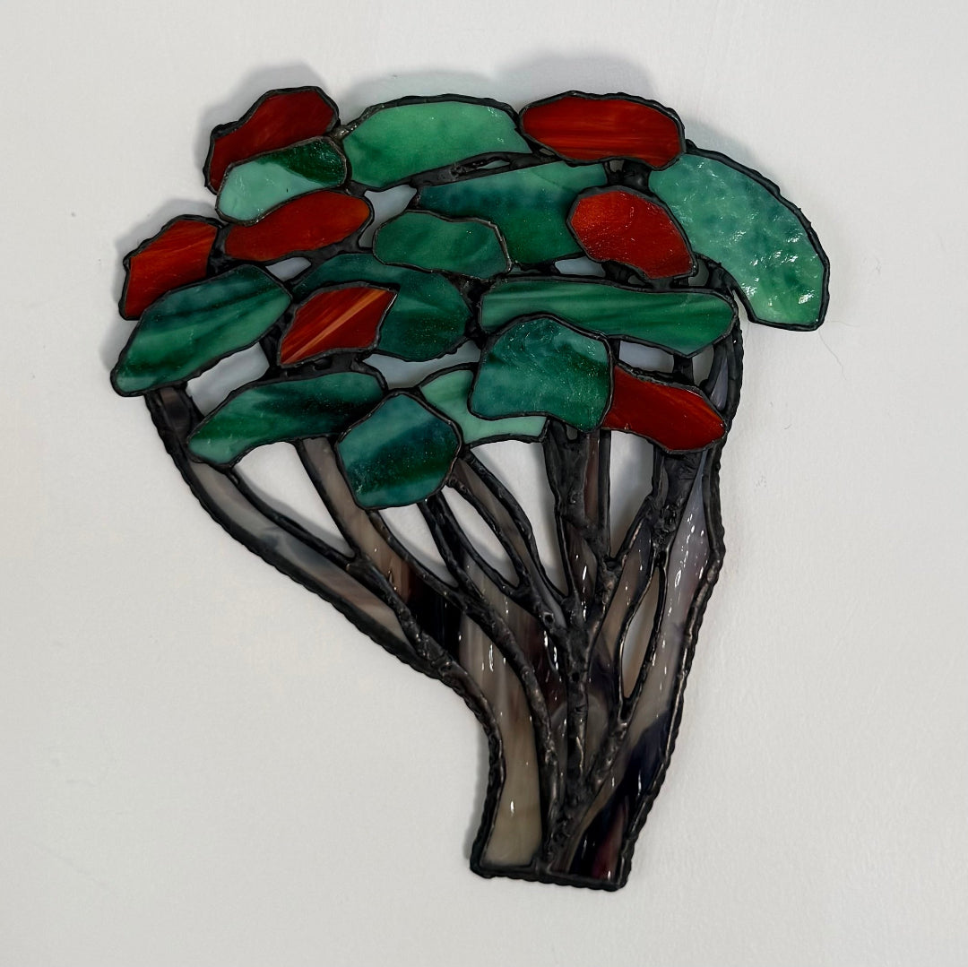 A stained glass art piece depicting a stylized tree with green leaves and red-orange fruit, set against a plain white background.