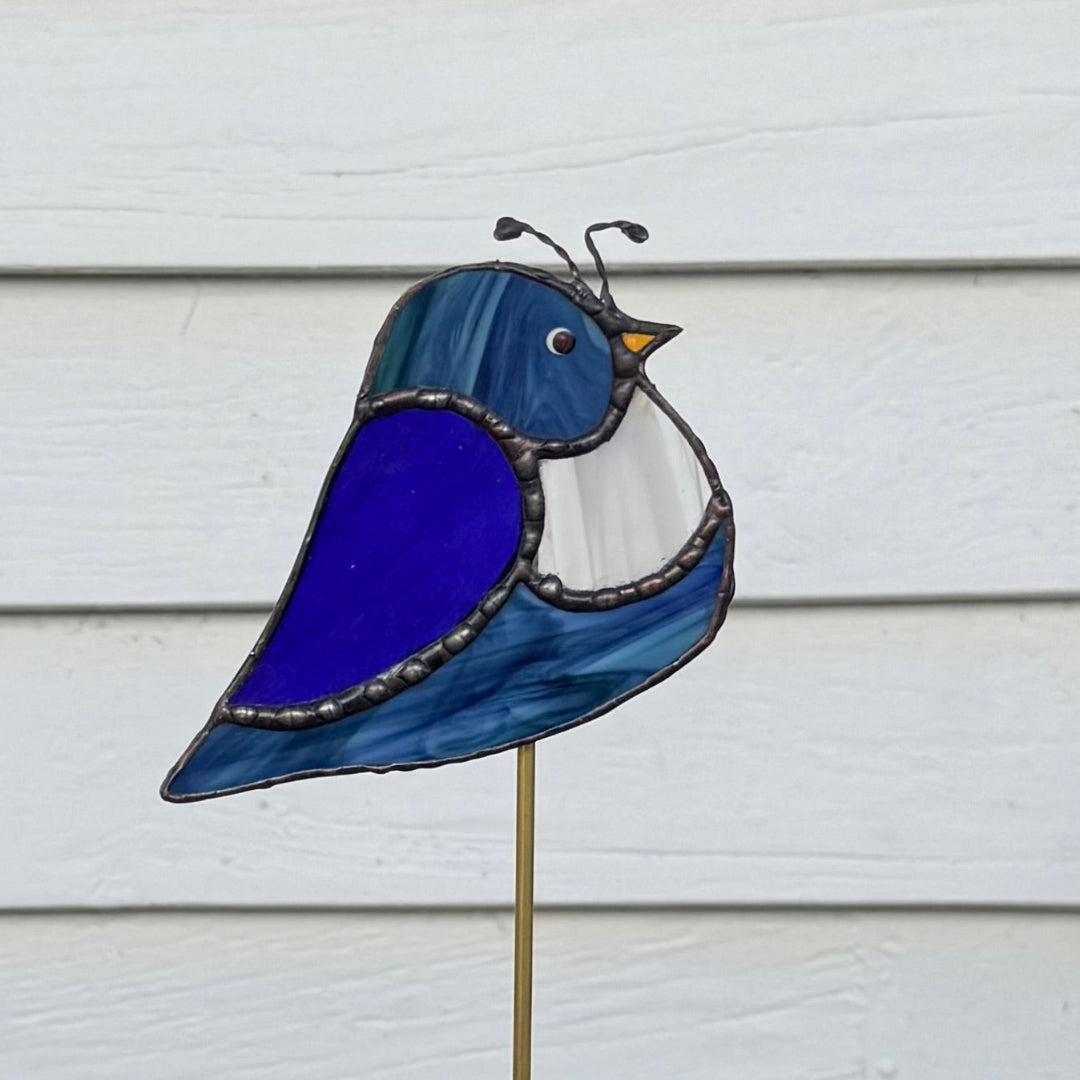 A stained glass garden stake shaped like a bird, featuring blue, purple, and white glass panels with dark metal outlines, mounted on a thin brass-colored stake against a white siding background.
