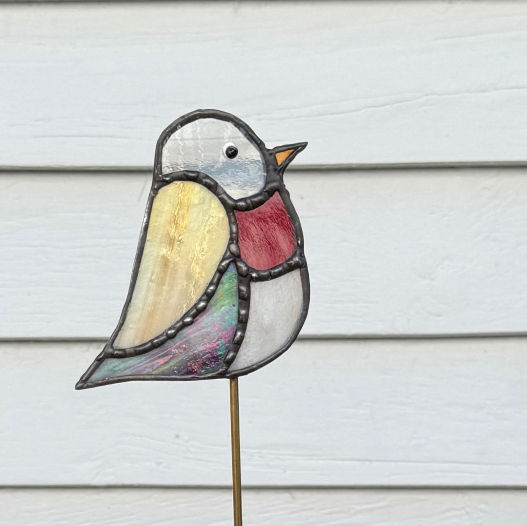 A stained glass bird garden stake featuring a white head, yellow wing, red chest, and iridescent green and purple tail, mounted on a thin metal stake against a white wood siding background.