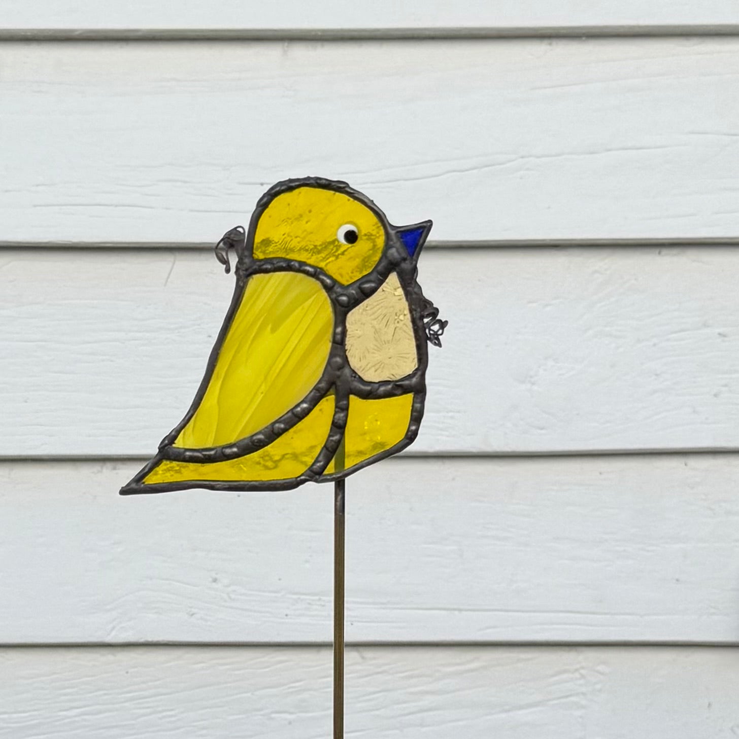 A stained glass bird garden stake featuring a yellow bird with a blue beak and cream chest, outlined in dark metal, mounted on a thin metal stake against a white wood siding background.