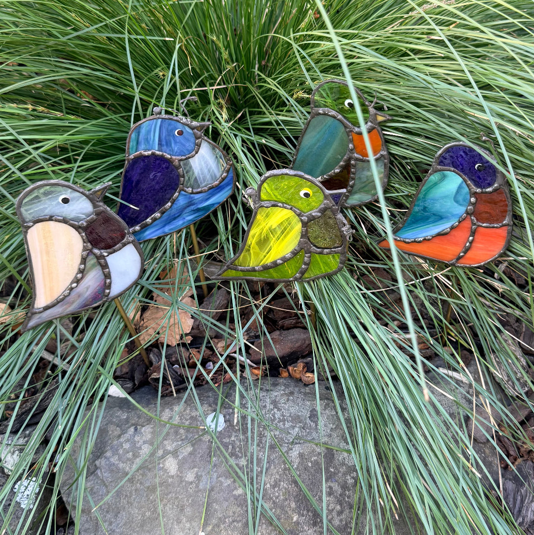Three colorful stained-glass bird garden stakes are displayed outdoors among green ornamental grass and a stone surface.