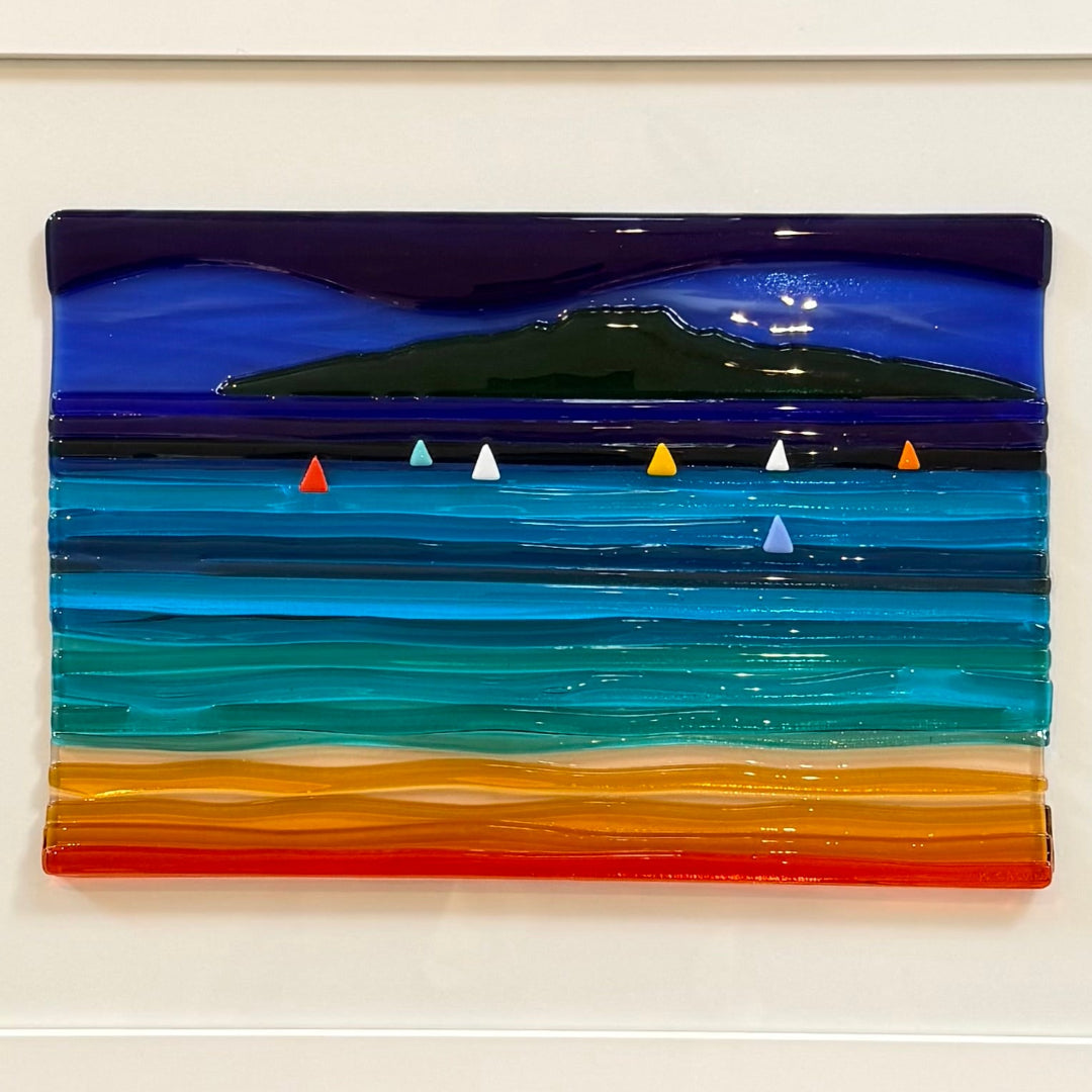 A square fused glass art piece depicting a coastal landscape with a dark green island under a deep blue night sky, blue and turquoise ocean waves, small colorful sailboats, and a sandy shore in orange and yellow tones, mounted on a white wall.