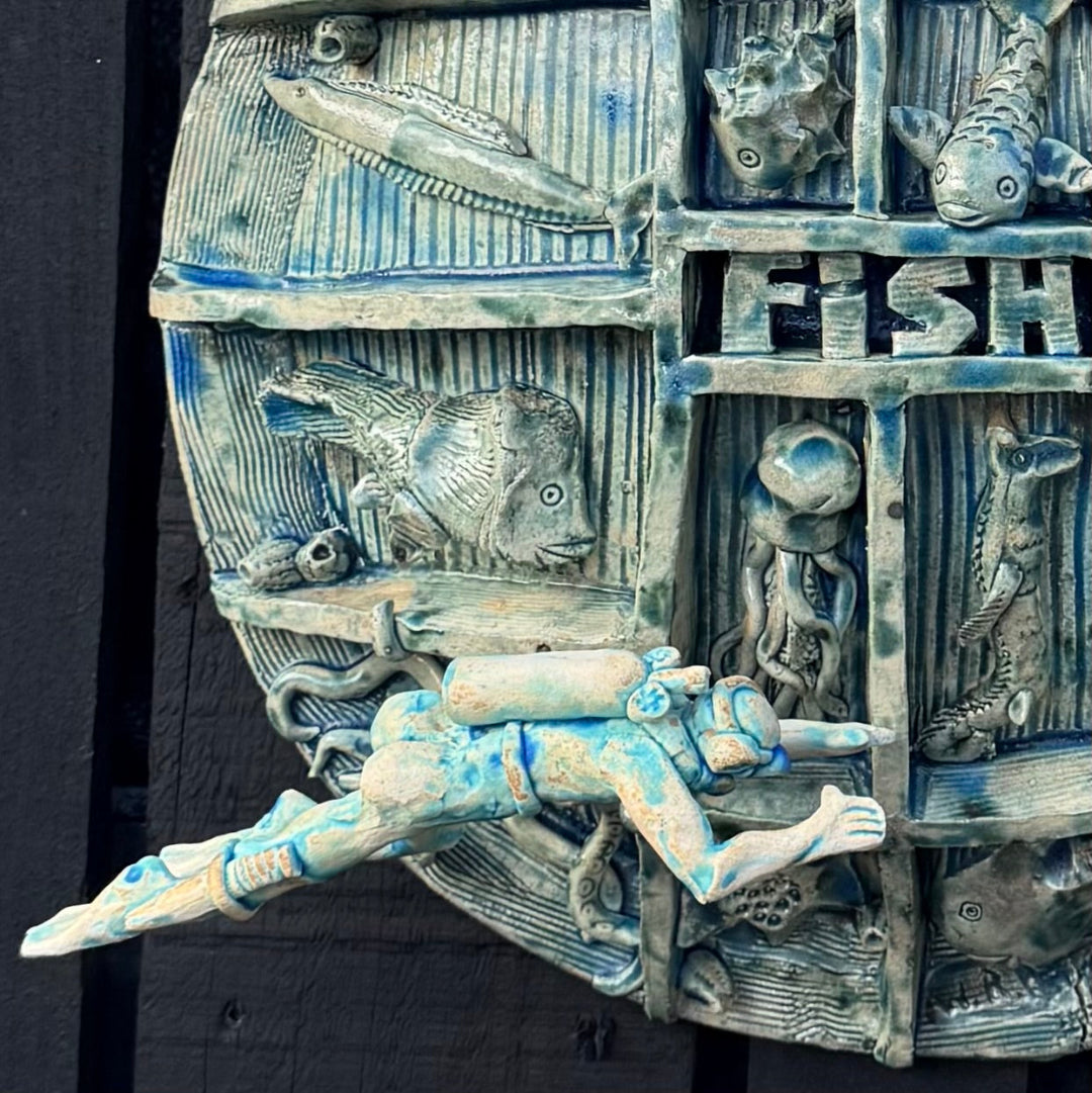 A close-up of a textured blue and gray ceramic wall sculpture featuring fish, an octopus, a diver, and the letters 'FISH' carved into a panel.